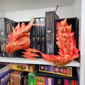 May include: A shelf displays fantasy-themed decor and books. A 3D-printed orange and gold dragon head and wings are featured. Books by Rebecca Yarros and J.R.R. Tolkien are visible, including "Fourth Wing" and "Onyx Storm."