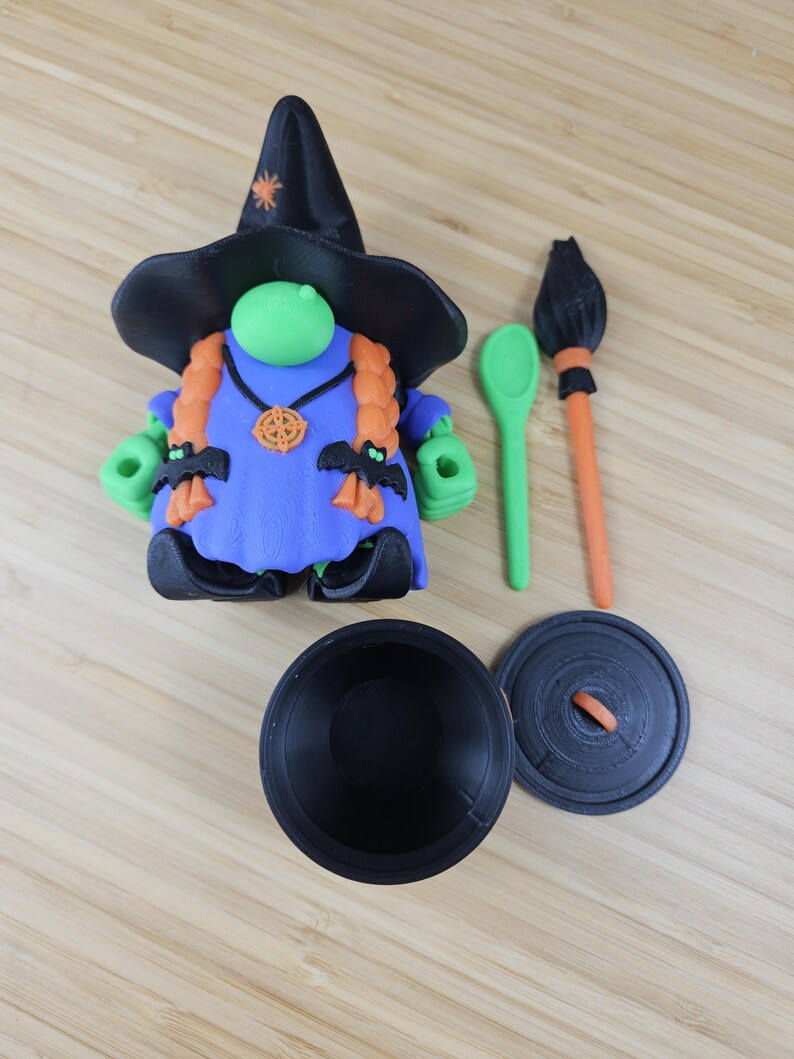 Puede incluir: Una figura caprichosa con tem&aacute;tica de Halloween de una bruja con sombrero negro y vestido azul. La bruja tiene trenzas naranjas, manos verdes y adornos en forma de murci&eacute;lago. Incluye un caldero, cuchara y escoba.