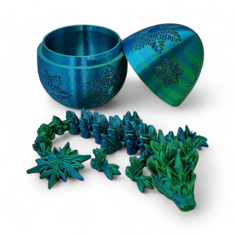 K&ouml;nnte beinhalten: Ein einzigartiges und fantasievolles Weihnachtsornament in Form eines Drachen, 3D-gedruckt in Tealblau und Gr&uuml;n mit Schneeflocken-Details. Das Ornament besteht aus zwei Teilen, einer hohlen Kugel und einer separaten Drachenfigur.