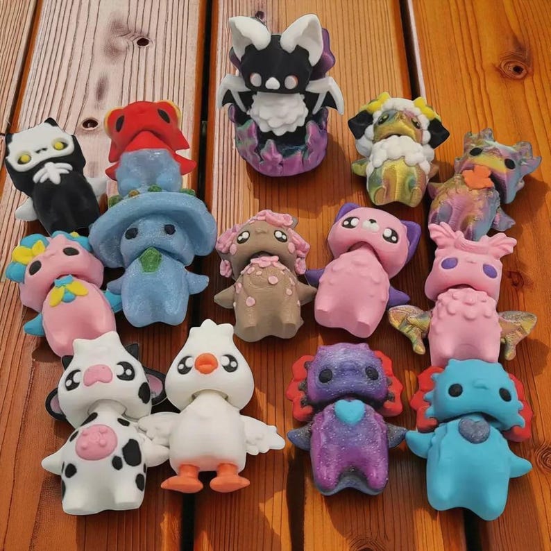 May include: A collection of colorful, whimsical figurines in various shapes and designs. The figurines include creatures with bat-like wings, cow-like spots, and bird-like features. The color palette includes pink, blue, purple, black, and white, set against a wooden surface.