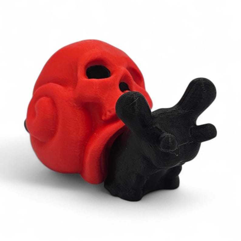 May include: A red skull-shaped ornament with a black hand-like figure emerging from its mouth. The skull has eye sockets and a nose cavity. The black figure has a hand shape with fingers extended. The objects are set against a white background.
