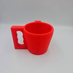 Pu&ograve; includere: Una tazza rosso vivo con superficie strutturata. Ha un manico rettangolare con apertura curva e una piccola cresta sul bordo. Progettata per bevande.