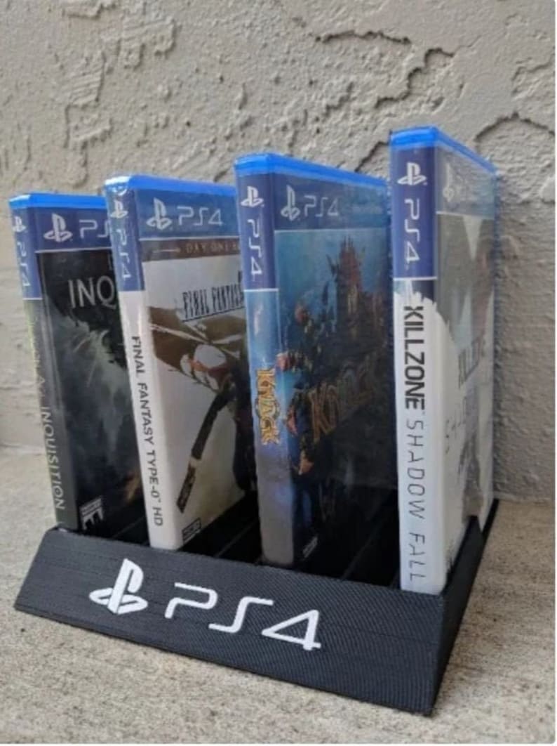 May include: A black plastic stand for storing video game cases. The stand has a PlayStation 4 logo on the front and can hold four game cases. The game cases are for the games Killzone: Shadow Fall, Final Fantasy Type-0 HD, Kingdom Hearts, and Dragon Age: Inquisition.