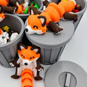 May include: 3D-printed articulated fox figurines in orange, white, and brown, posed near and on top of gray trash cans. The foxes have a segmented body and tail, with a playful, cartoonish design. The image is set against a white background.