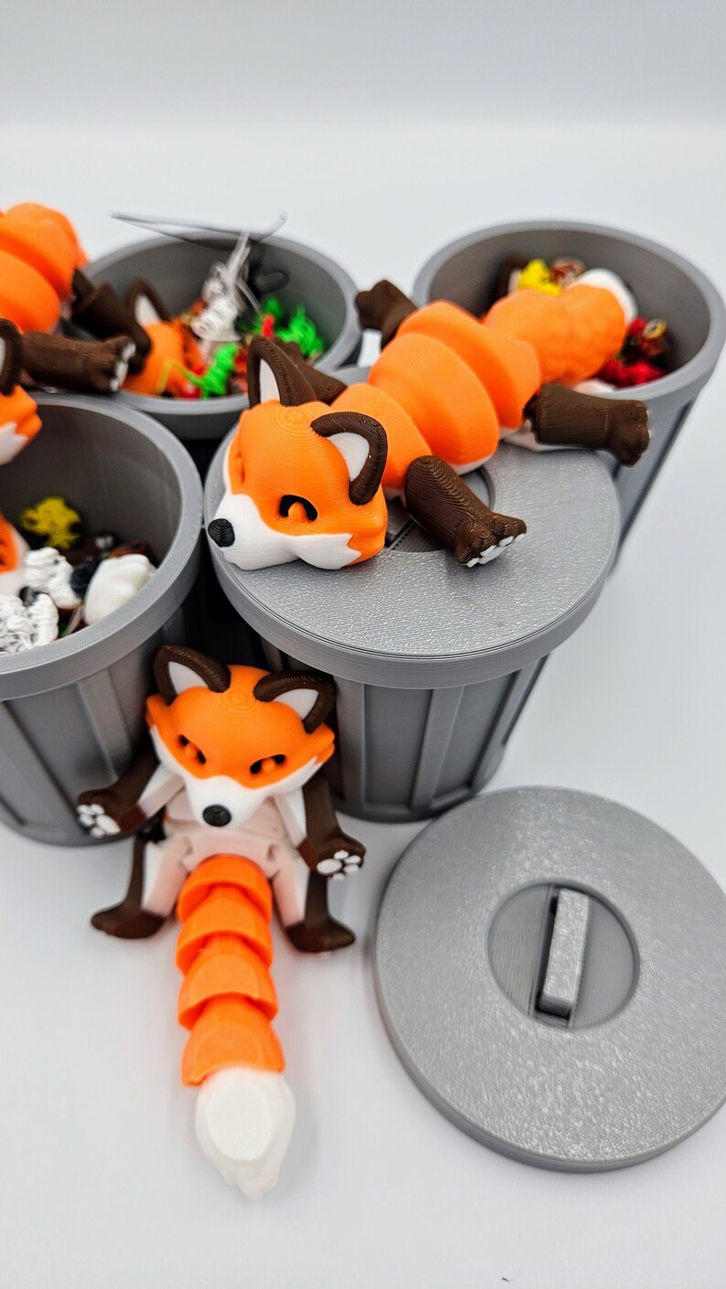 May include: 3D-printed articulated fox figurines in orange, white, and brown, posed near and on top of gray trash cans. The foxes have a segmented body and tail, with a playful, cartoonish design. The image is set against a white background.