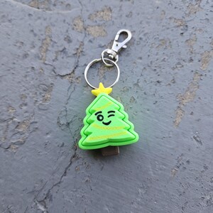 Pode incluir: Um chaveiro de &aacute;rvore de Natal verde com uma cara piscando e uma estrela amarela no topo. O chaveiro tem um anel de metal prateado e um fecho de metal prateado.