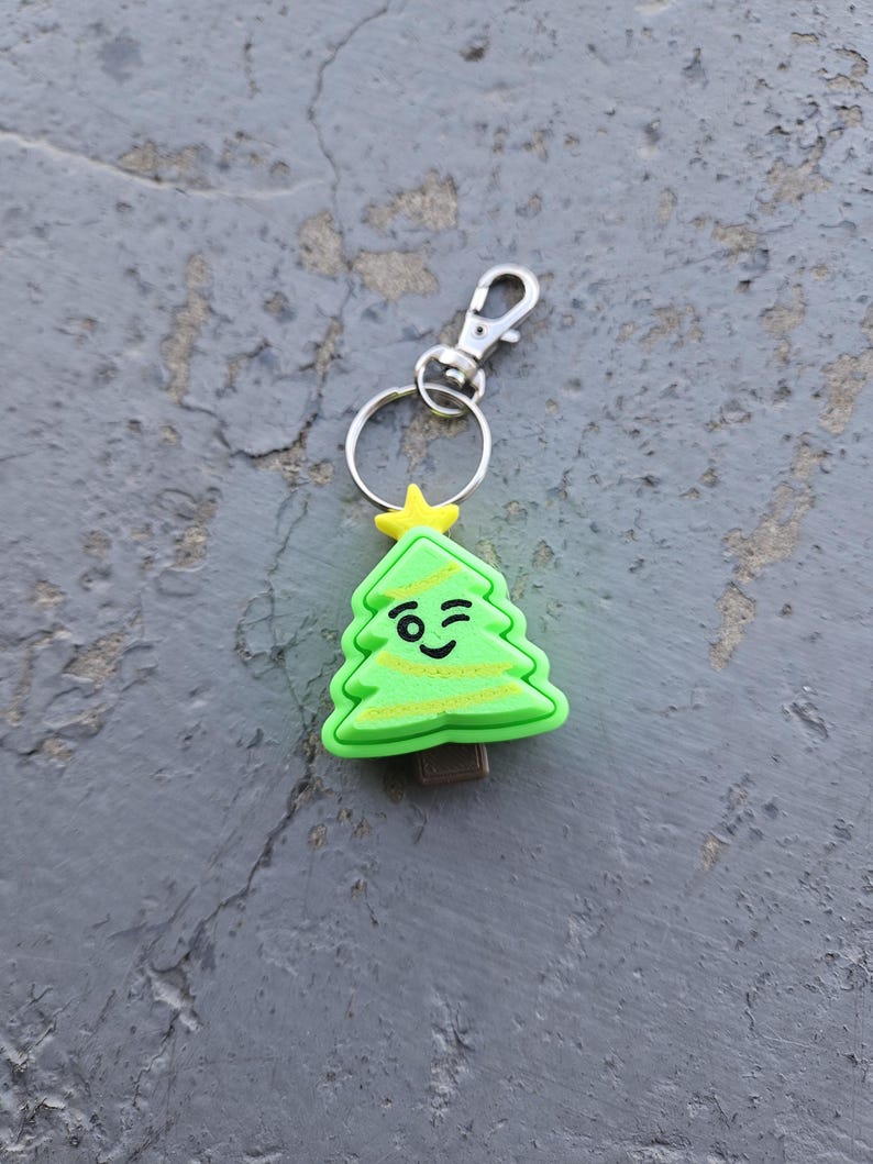 Pode incluir: Um chaveiro de &aacute;rvore de Natal verde com uma cara piscando e uma estrela amarela no topo. O chaveiro tem um anel de metal prateado e um fecho de metal prateado.