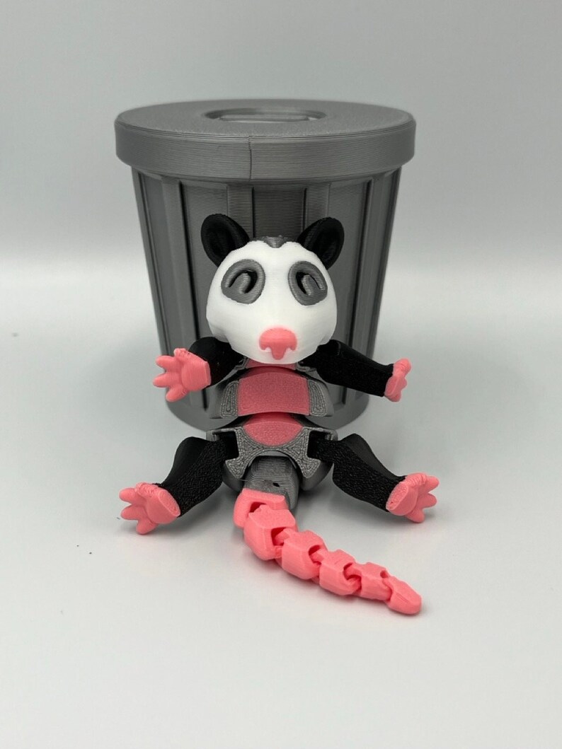 Op de afbeelding: Een 3D-geprint opossum-speelgoed met een roze staart, voeten en neus, zwarte armen en oren en een wit gezicht. Het speelgoed staat voor een grijze vuilnisbak. Het lichaam van de opossum is grijs en roze.