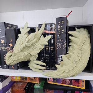 Op de afbeelding: Een paar lichtgroene boekensteunen in de vorm van een draak. E&eacute;n is de kop, de ander de staart. Ze staan op een witte plank tussen boeken. Decoratieve boekensteunen voor liefhebbers van fantasy.