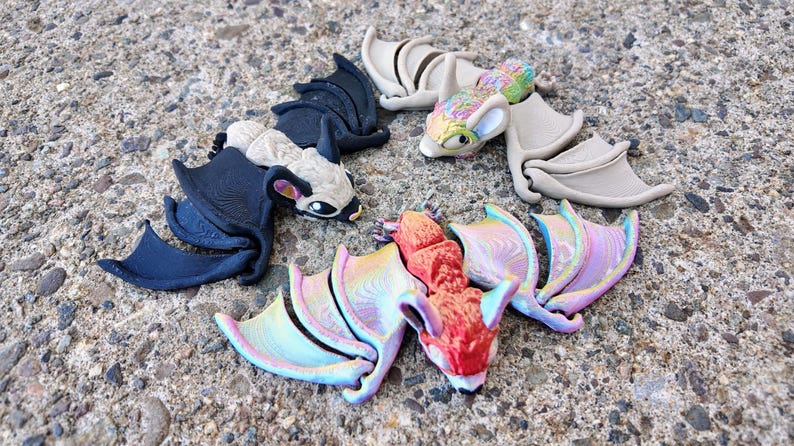 May include: Four colorful bat figurines with wings outstretched. The bats are made of a smooth, textured material and are painted in various shades of black, white, gray, red, pink, and blue.