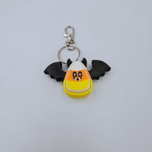 May include: A black bat-shaped keychain with a candy corn design. The candy corn is white, orange, and yellow with a black face.