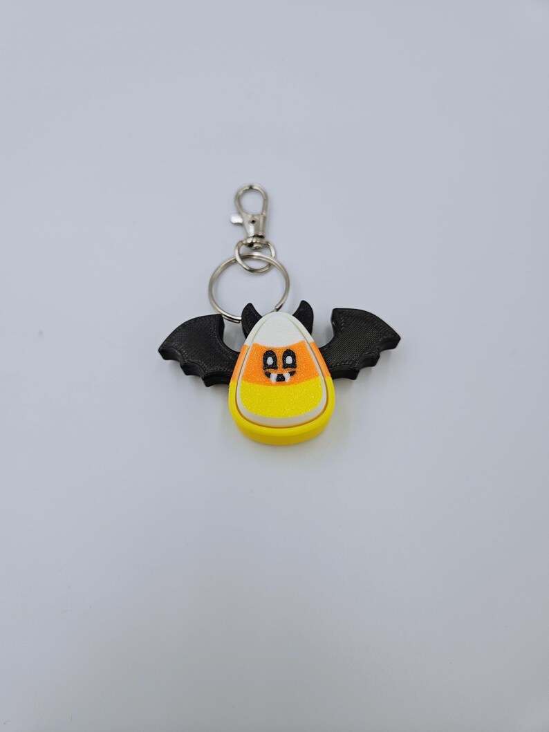 May include: A black bat-shaped keychain with a candy corn design. The candy corn is white, orange, and yellow with a black face.