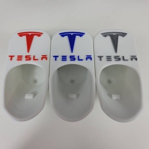 May include: Three white 3D printed wall mounts shaped like Tesla car charging ports. The mounts have the Tesla logo and the word "TESLA" printed on them. The logos are in red, blue, and gray.