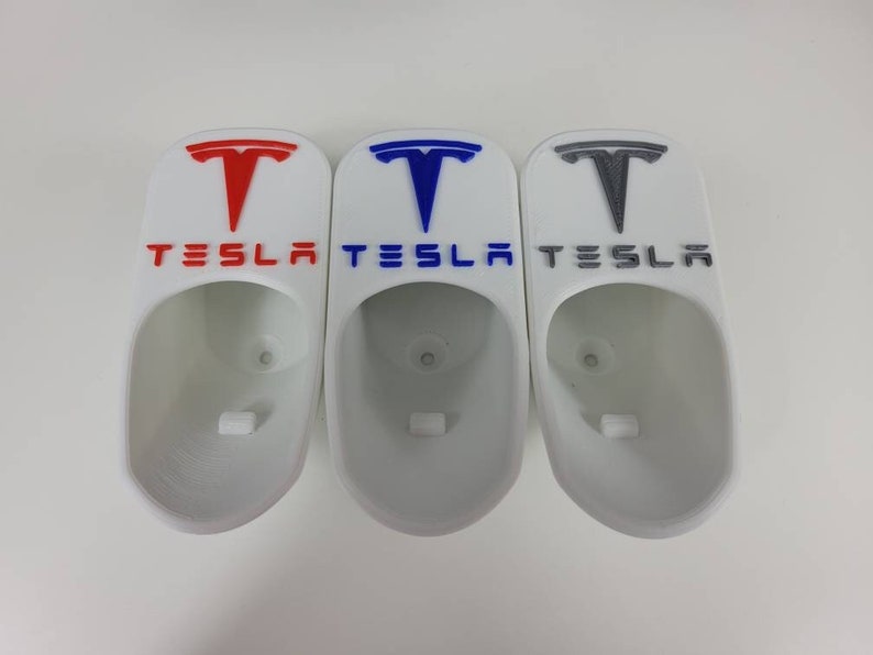 May include: Three white 3D printed wall mounts shaped like Tesla car charging ports. The mounts have the Tesla logo and the word "TESLA" printed on them. The logos are in red, blue, and gray.