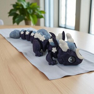 May include: A dark blue dragon figurine with light blue and white accents. The dragon has a segmented body, with a head and tail. The dragon is lying on a light blue cloth on a wooden table. The dragon's design includes snowflake details.