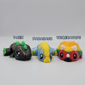 May include: Three small dinosaur figurines, painted in different colors. The T-Rex is black with green spikes, the Parasaur is blue with a yellow crest, and the Triceratops is red, yellow, and green.