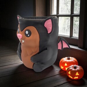May include: A Halloween-themed ceramic container shaped like a cartoon bat with a brown body, black bat wings, and pink accents. Two small, orange jack-o'-lanterns with lit faces sit nearby on a wooden surface, near a window.