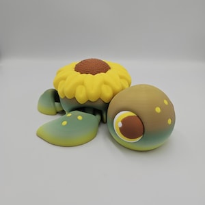 Pode incluir: Uma figura de tartaruga caprichosa impressa em 3D com uma concha de girassol. A tartaruga apresenta um topo de girassol amarelo com um centro marrom, barbatanas verdes e amarelas e uma cabe&ccedil;a marrom e verde com um olho grande e detalhado.