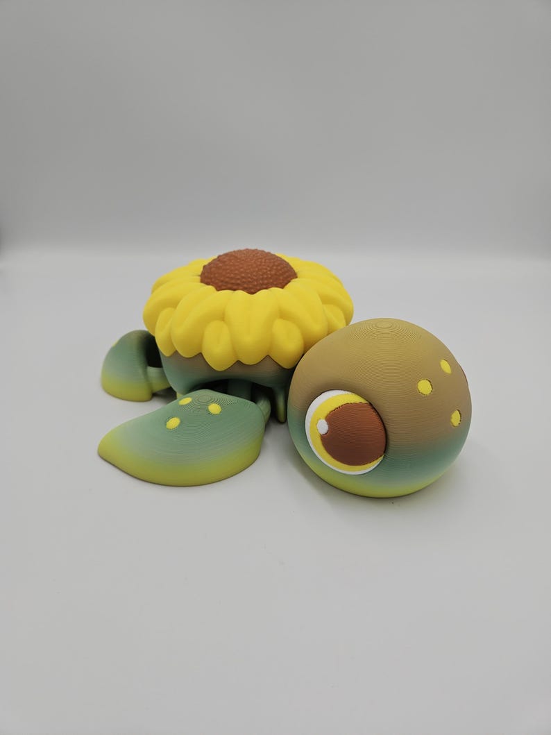 Pode incluir: Uma figura de tartaruga caprichosa impressa em 3D com uma concha de girassol. A tartaruga apresenta um topo de girassol amarelo com um centro marrom, barbatanas verdes e amarelas e uma cabe&ccedil;a marrom e verde com um olho grande e detalhado.