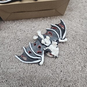 May include: Black and white bat-shaped decorative objects with red accents. The bats have a skeletal design with open wings and are lying on a gray carpet. Some bats are near a brown cardboard box.