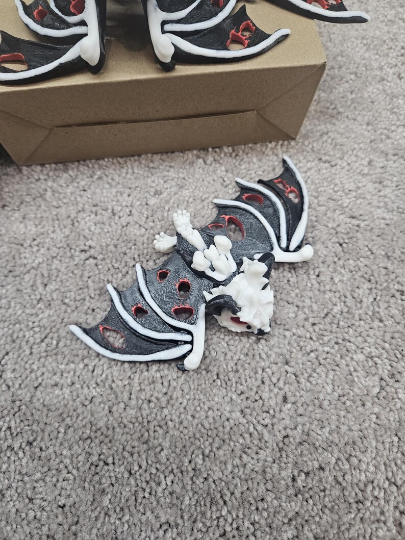 May include: Black and white bat-shaped decorative objects with red accents. The bats have a skeletal design with open wings and are lying on a gray carpet. Some bats are near a brown cardboard box.