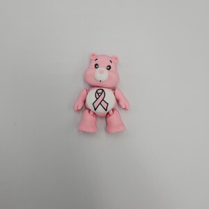 May include: A pink bear figurine with a white belly featuring a pink ribbon symbol. The bear has black eyes, a white nose, and is posed against a plain white background. The figurine is a representation of a Care Bear.