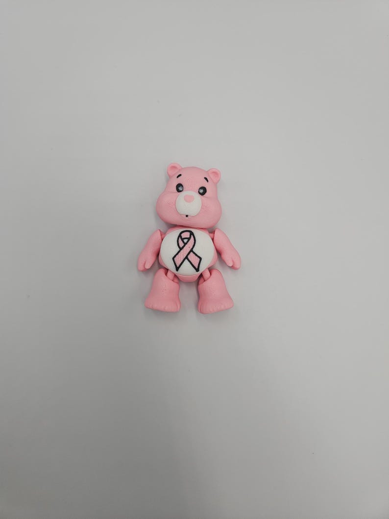 May include: A pink bear figurine with a white belly featuring a pink ribbon symbol. The bear has black eyes, a white nose, and is posed against a plain white background. The figurine is a representation of a Care Bear.