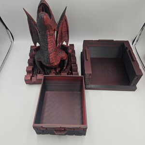 May include: A red and black dragon sculpture with open mouth and wings, atop a brick-like base. Two matching red boxes are also present, one open and one closed, suggesting a storage or display theme.