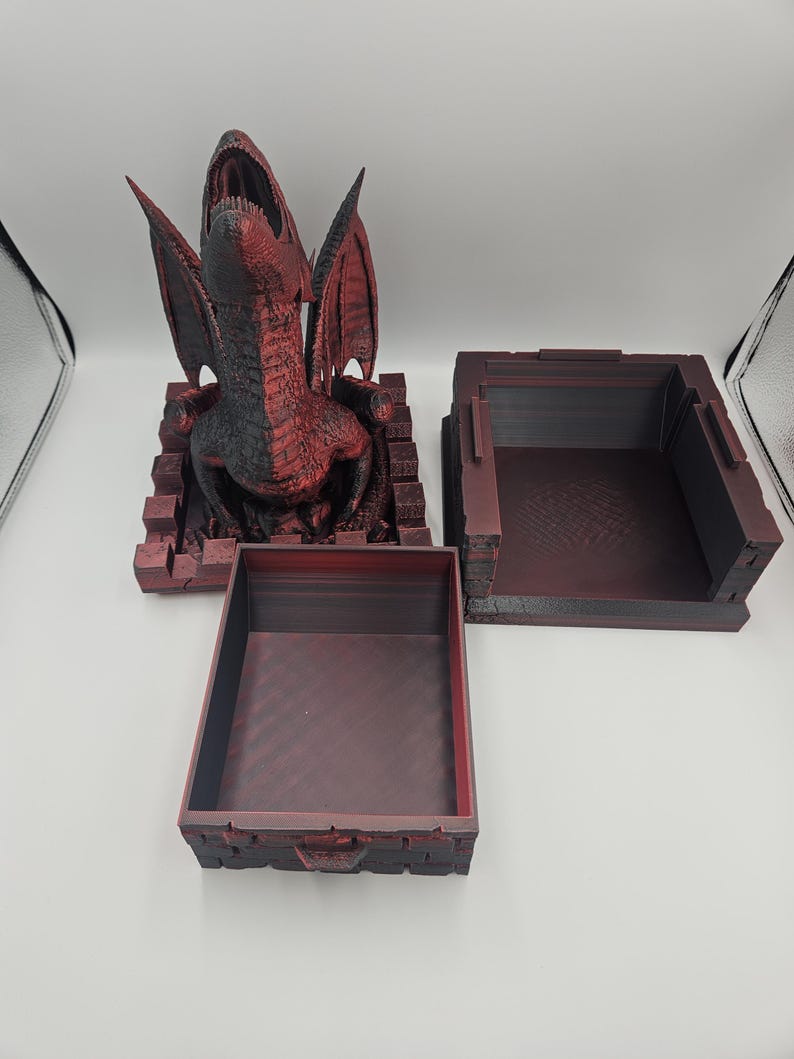 May include: A red and black dragon sculpture with open mouth and wings, atop a brick-like base. Two matching red boxes are also present, one open and one closed, suggesting a storage or display theme.