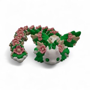 May include: A whimsical, handcrafted sculpture of a creature with a white head, green eyes, and a body adorned with pink flowers and green leaves. The sculpture is made of a light-colored material and sits on a white surface.