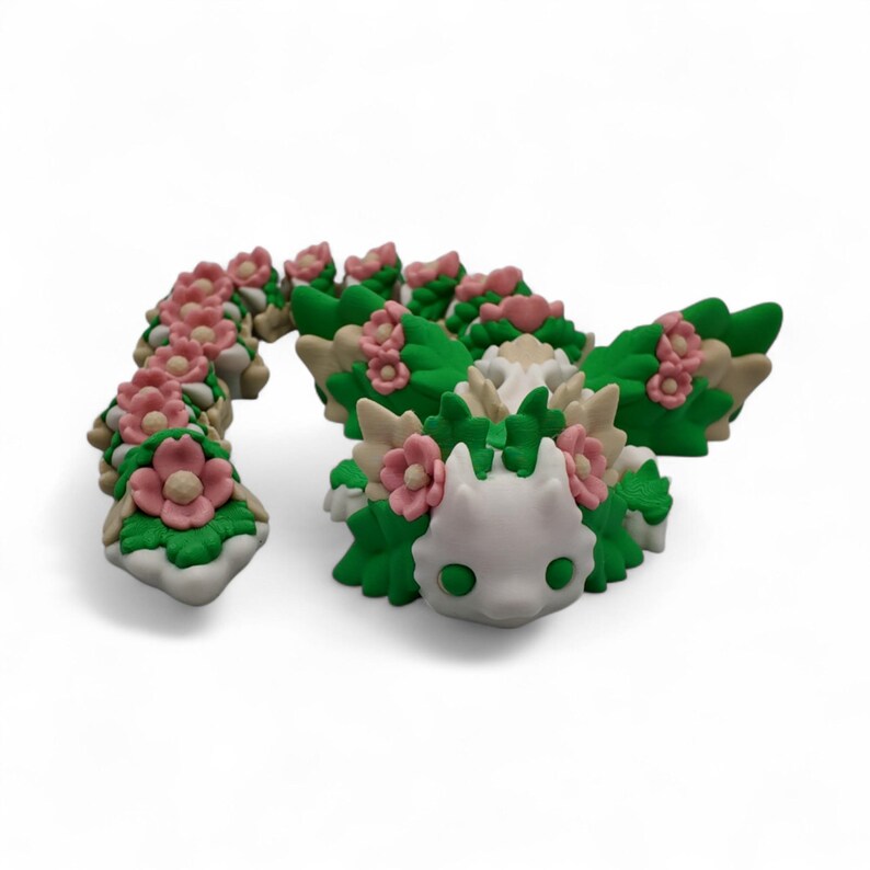 May include: A whimsical, handcrafted sculpture of a creature with a white head, green eyes, and a body adorned with pink flowers and green leaves. The sculpture is made of a light-colored material and sits on a white surface.