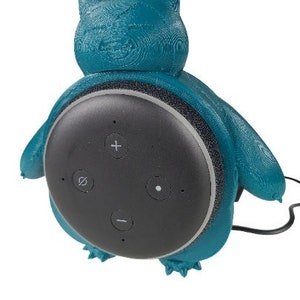 May include: A teal blue 3D printed  speaker stand shaped like a cartoon character. The stand is designed to hold a round, black smart speaker with four buttons.