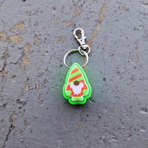 Pode incluir: Um chaveiro de gnomo verde com um chap&eacute;u listrado vermelho e branco. O gnomo tem uma barba castanha e um rosto branco. O chaveiro tem um anel de metal prateado e um fecho de metal prateado.