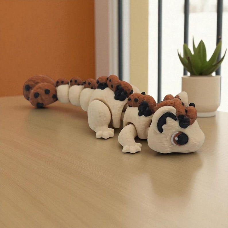 May include: An articulated toy creature with a segmented body, primarily off-white with brown and black details. The toy is designed to resemble a caterpillar or a stylized animal. It is positioned on a light-coloured wooden surface.