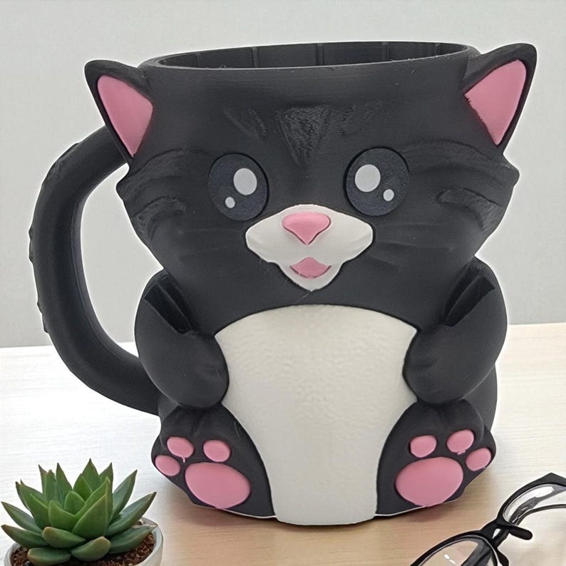 May include: A black cat-shaped mug with pink inner ears, nose, and paw pads. The mug has a white chest and face, with large, round eyes. The handle is a curved tail. A fun and whimsical item for cat lovers.