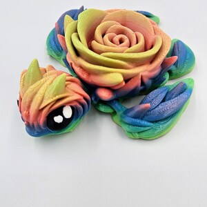 May include: A 3D printed rainbow colored rose with a smaller rose head attached to the side. The rose is made of plastic and has a detailed, textured surface.