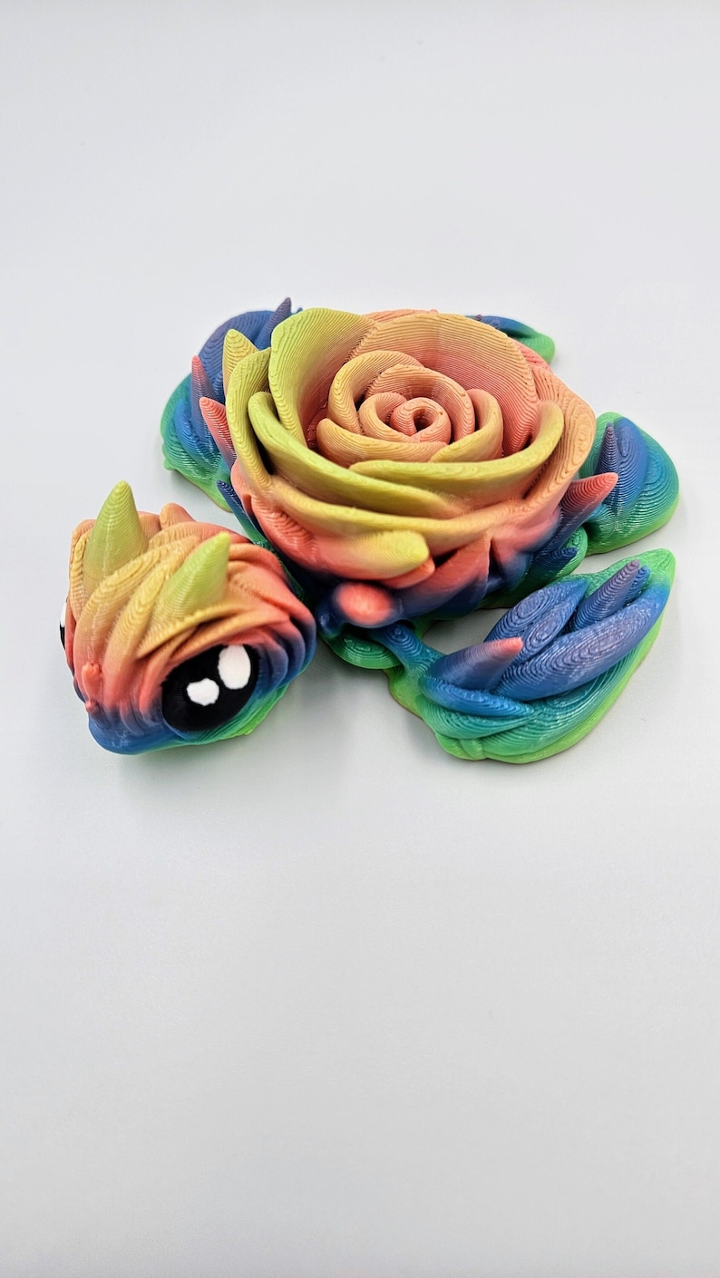 May include: A 3D printed rainbow colored rose with a smaller rose head attached to the side. The rose is made of plastic and has a detailed, textured surface.