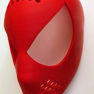 May include: A bright red mask with a smooth finish, featuring a large eye opening and a perforated top section. The mask is designed with a curved shape and a prominent nose area, suggesting a superhero or character theme.
