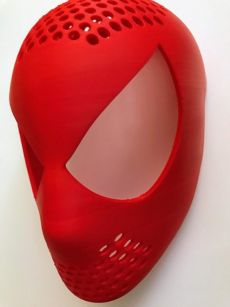 May include: A bright red mask with a smooth finish, featuring a large eye opening and a perforated top section. The mask is designed with a curved shape and a prominent nose area, suggesting a superhero or character theme.