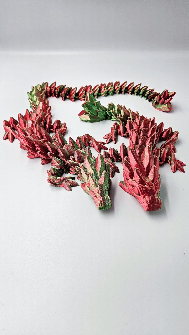 K&ouml;nnte beinhalten: Ein 3D-gedruckter Drache aus rotem und gr&uuml;nem Kunststoff. Der Drache ist zu einem Kreis zusammengerollt, wobei sich Kopf und Schwanz ber&uuml;hren. Der Drache besteht aus vielen kleinen, scharfen Schuppen.