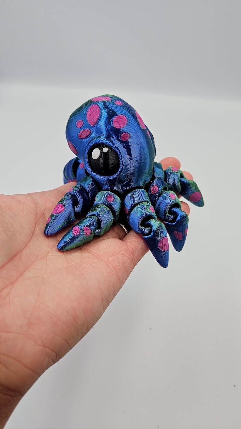 Puede incluir: Un peque&ntilde;o pulpo azul y verde con lunares rosas. El pulpo tiene un ojo grande y negro y est&aacute; hecho de pl&aacute;stico impreso en 3D.