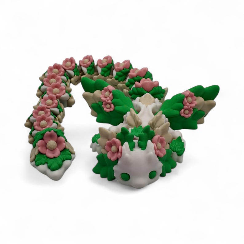 May include: A whimsical, handcrafted sculpture of a dragon, featuring a white body, green wings, and a tail. The dragon is adorned with pink flowers and green leaves, creating a fantasy-themed aesthetic. The sculpture is set against a white background.