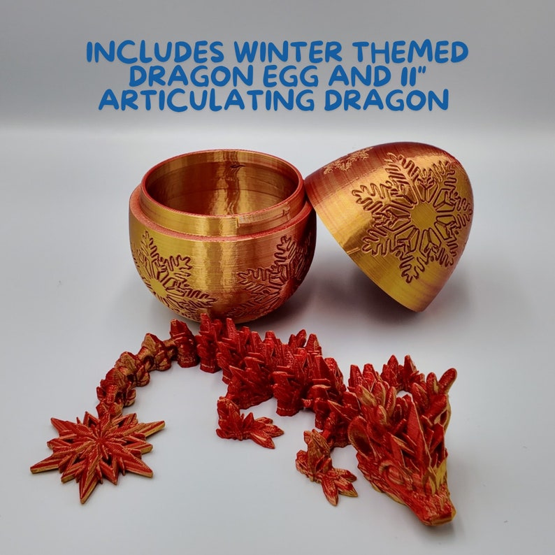 K&ouml;nnte beinhalten: Ein goldenes und rotes Dracheneier-Ornament mit Schneeflockenmuster. Das Ei ist ge&ouml;ffnet und enth&uuml;llt einen roten, artikulierten Drachen im Inneren. Enth&auml;lt ein winterliches Dracheneier-Ornament und einen 28 cm langen, artikulierten Drachen.