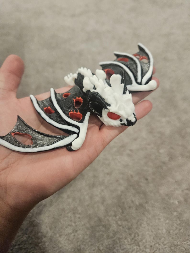 May include: A pair of dragon figurines, each featuring a black body with red accents and white detailing. The dragons have red eyes and are held in a hand. The wings are black with a silver outline.