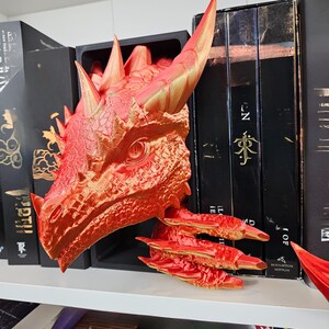 May include: A red and gold dragon head bookend, with detailed scales and horns. The dragon's head is positioned to appear as if it is emerging from a shelf, with its claws extending out. The bookend is placed among a collection of black books.