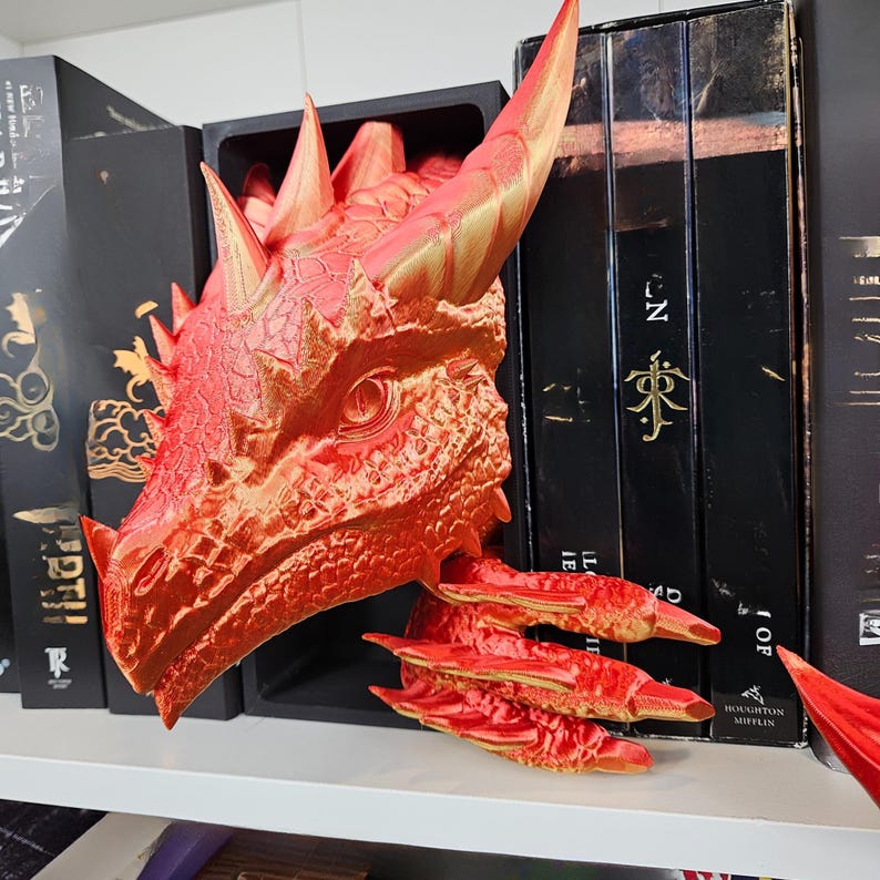 May include: A red and gold dragon head bookend, with detailed scales and horns. The dragon's head is positioned to appear as if it is emerging from a shelf, with its claws extending out. The bookend is placed among a collection of black books.