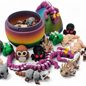 K&ouml;nnte beinhalten: Ein regenbogenfarbener eif&ouml;rmiger Beh&auml;lter mit Deckel, gef&uuml;llt mit kleinen Tierfiguren. Die Figuren umfassen einen lila Drachen, einen gr&uuml;nen Dinosaurier, eine braune Eule, einen grauen Elefanten, einen grauen Opossum, einen braun-wei&szlig;en Axolotl, einen grau-wei&szlig;en Opossum, einen gr&uuml;nen Frosch, eine braun-wei&szlig;e Eidechse, einen lila segmentierten Drachen, eine schwarze Spinne und eine beige Eidechse.