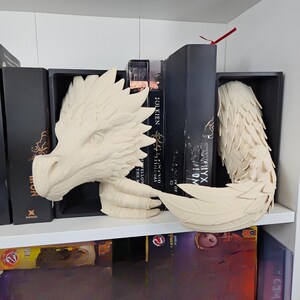 May include: A cream-colored dragon head and tail bookends. The dragon head features detailed scales and a sharp, pointed crest. The tail is curled and has a feathered appearance. The bookends are displayed on a white shelf with books.