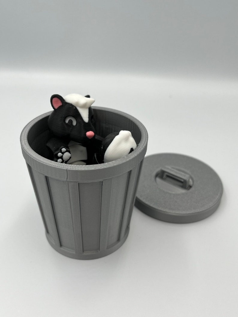 May include: A gray 3D-printed trash can with a removable lid. Inside the can is a black and white skunk figurine. The skunk is in a sitting position. The skunk has a white stripe down its back and a white patch on its head.