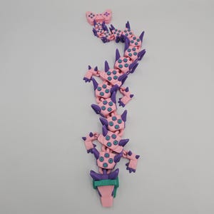 May include: A pink and purple articulated dragon toy with teal accents. The dragon has a controller at the top and a green and pink base. The toy is made of plastic and is designed to be flexible and poseable.